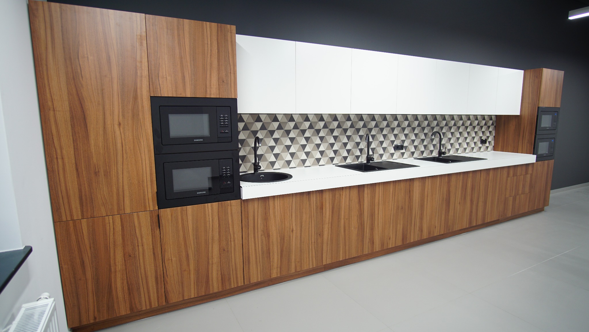 Nowoczesna kuchnia z drewnianymi frontami, białymi szafkami górnymi i czarnymi detalami. Dwie czarne mikrofalówki w zabudowie. Geometryczny wzór na ścianie nad blatem.