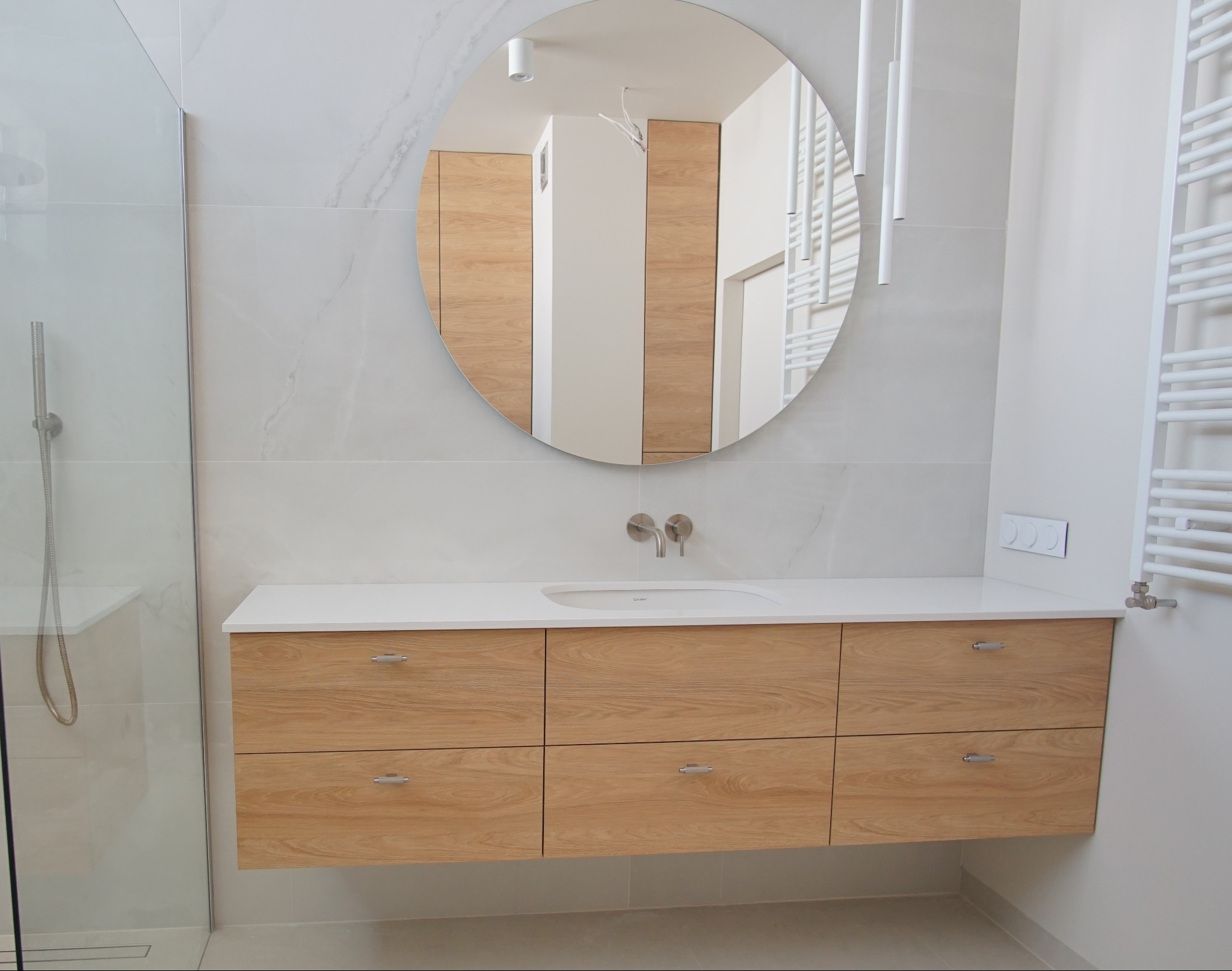 Nowoczesna łazienka z drewnianą szafką pod umywalką i okrągłym lustrem, minimalistyczny design, białe płytki imitujące marmur, grzejnik drabinkowy.