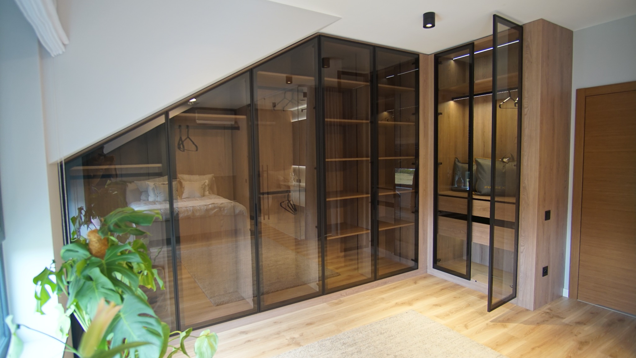 Nowoczesna szafa wnękowa z przesuwnymi, szklanymi drzwiami w aranżacji pokoju. Widoczne półki, wieszaki i elementy garderoby. Ujęcie z rośliną w rogu.