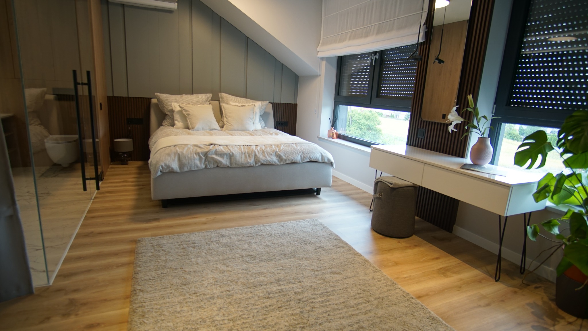 Nowoczesna sypialnia z łazienką en-suite. Jasne drewno na podłodze i ścianach, duże okna z roletami, minimalistyczne biurko i dekoracje roślinne tworzą przytulną atmosferę.