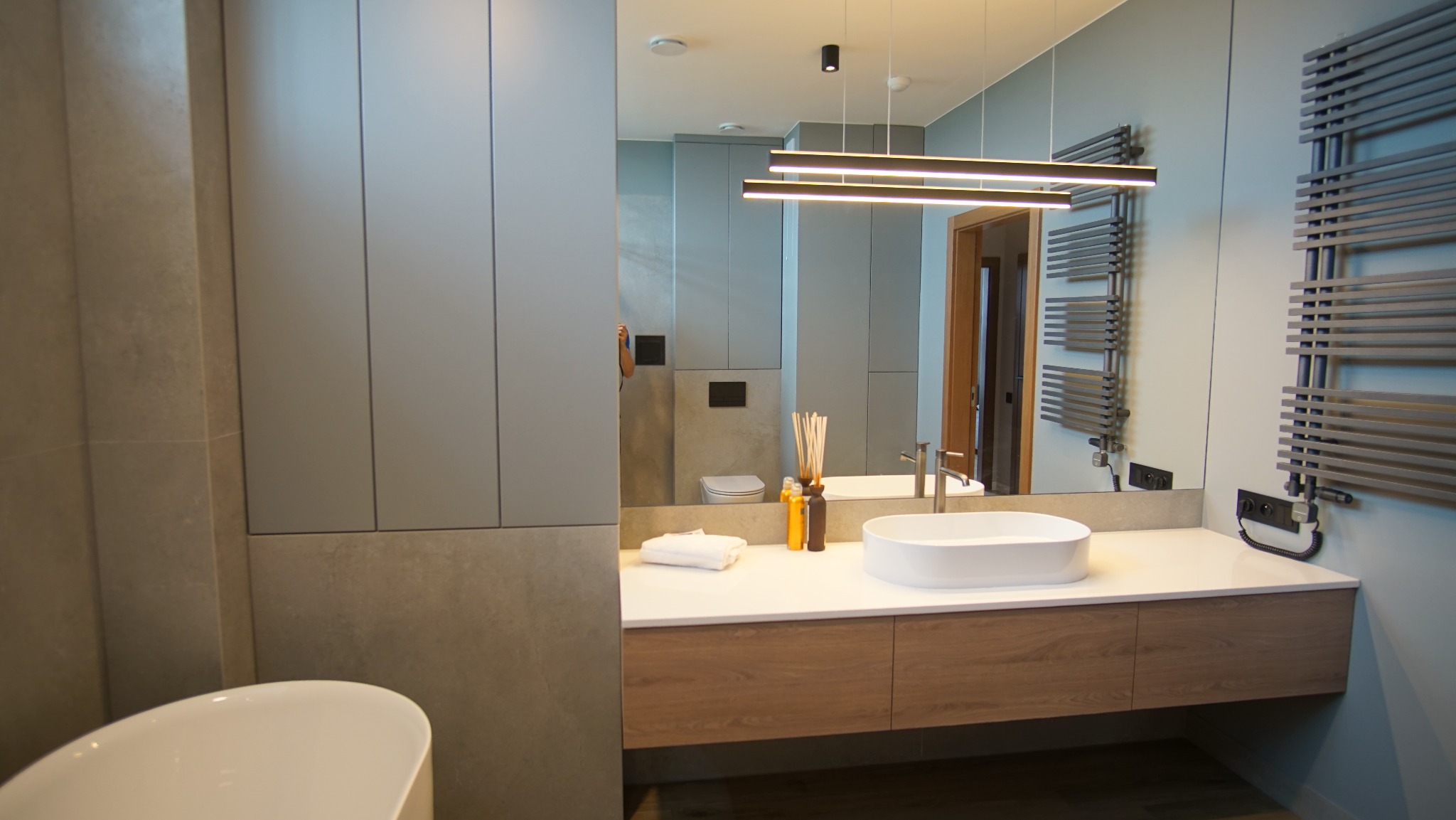 Nowoczesna łazienka z umywalką nablatową, drewnianą szafką pod umywalką i grzejnikiem drabinkowym. Duże lustro odbija dalszą część łazienki.