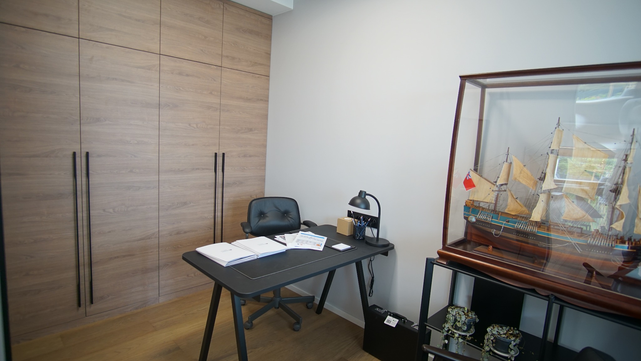 Biuro w domu z dużą drewnianą szafą wnękową, czarnym biurkiem i krzesłem. Na biurku dokumenty, lampka i model żaglowca w szklanej gablocie.