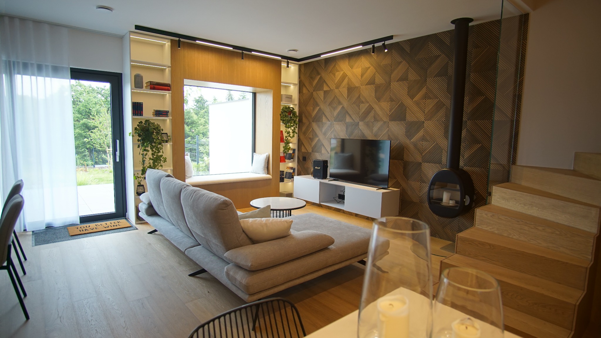 Nowoczesny salon z szarą sofą, oknem z siedziskiem, ścianą z geometrycznym wzorem i wiszącym kominkiem. Na podłodze drewniane schody. Całość w ciepłej tonacji.