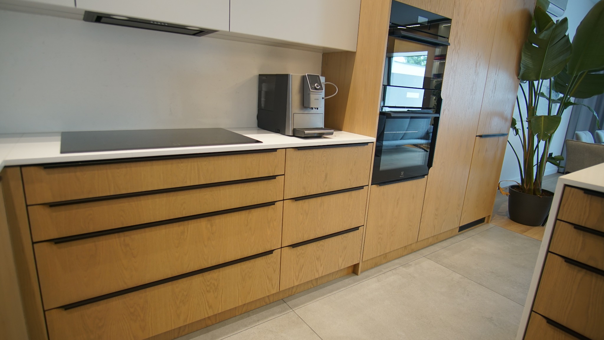 Nowoczesna kuchnia z drewnianymi frontami, czarnymi uchwytami i sprzętem AGD w zabudowie. Na blacie ekspres do kawy. Minimalistyczny design. Roślina doniczkowa w rogu.