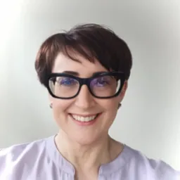 Portret uśmiechniętej kobiety w okularach o grubych oprawkach, ubrana w bluzkę w kolorze liliowym. Krótkie, ciemne włosy i jasna karnacja.