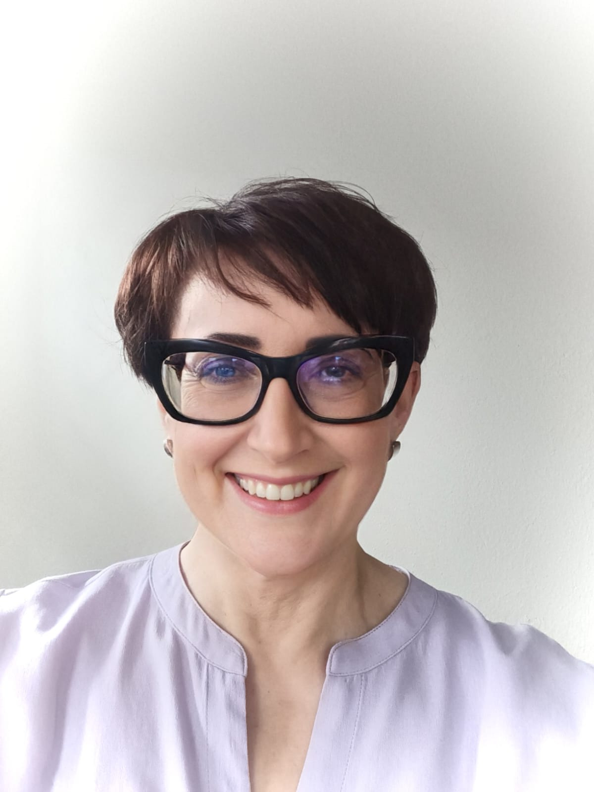 Portret uśmiechniętej kobiety w okularach o grubych oprawkach, ubrana w bluzkę w kolorze liliowym. Krótkie, ciemne włosy i jasna karnacja.