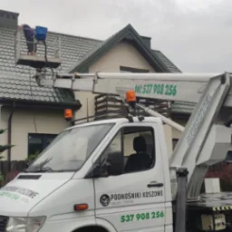Pracownik na podnośniku koszowym myje dach domu jednorodzinnego. Widoczny biały samochód z napisem 'Podnośniki Koszowe' i numerem telefonu. Pochmurne niebo w tle.
