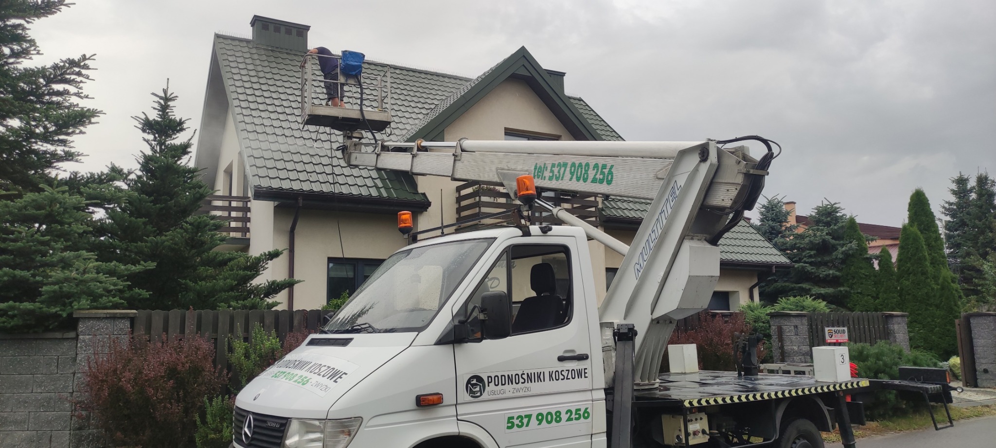 Pracownik na podnośniku koszowym myje dach domu jednorodzinnego. Widoczny biały samochód z napisem 'Podnośniki Koszowe' i numerem telefonu. Pochmurne niebo w tle.