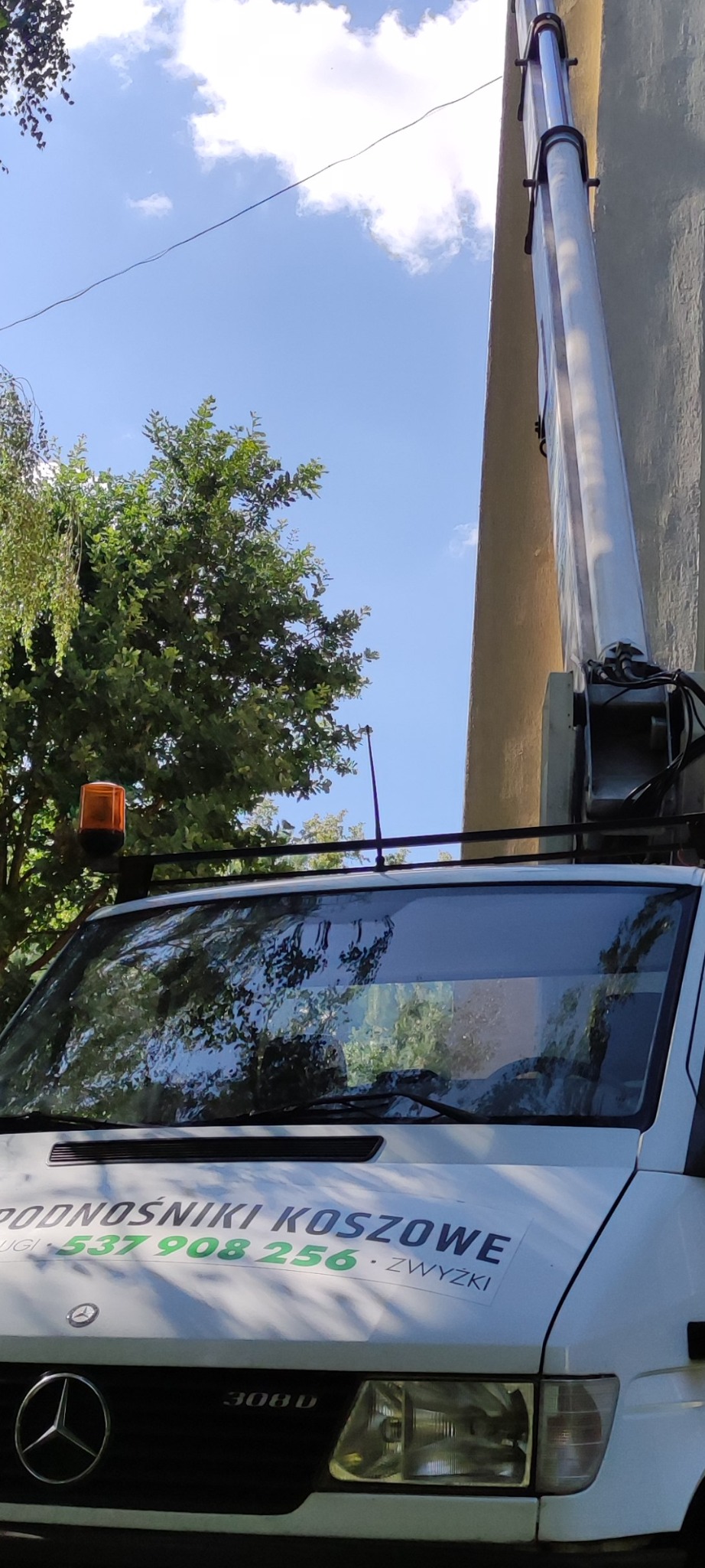 Podnośnik koszowy na tle błękitnego nieba i zieleni drzew, z wysuniętym ramieniem przy budynku. Widoczny fragment pojazdu z reklamą usług.
