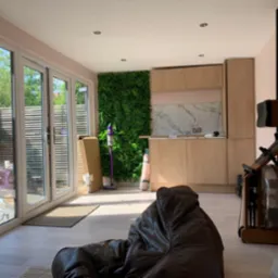 Jasne wnętrze z drewnianą zabudową kuchenną, ścianą z roślin, sprzętem do ćwiczeń i dużymi oknami balkonowymi. Widoczny fragment brązowego worka sako.