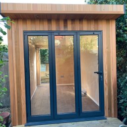 WoodMaster - Nowoczesna, drewniana altana z trzyskrzydłowymi, przeszklonymi drzwiami w grafitowej ramie, z widocznym wnętrzem i elementami wyposażenia.