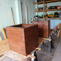 WoodMaster - Dwie drewniane, bejcowane donice w kształcie sześcianu, ustawione na kozłach warsztatowych w pomieszczeniu z półkami. Widoczne ślady pracy.