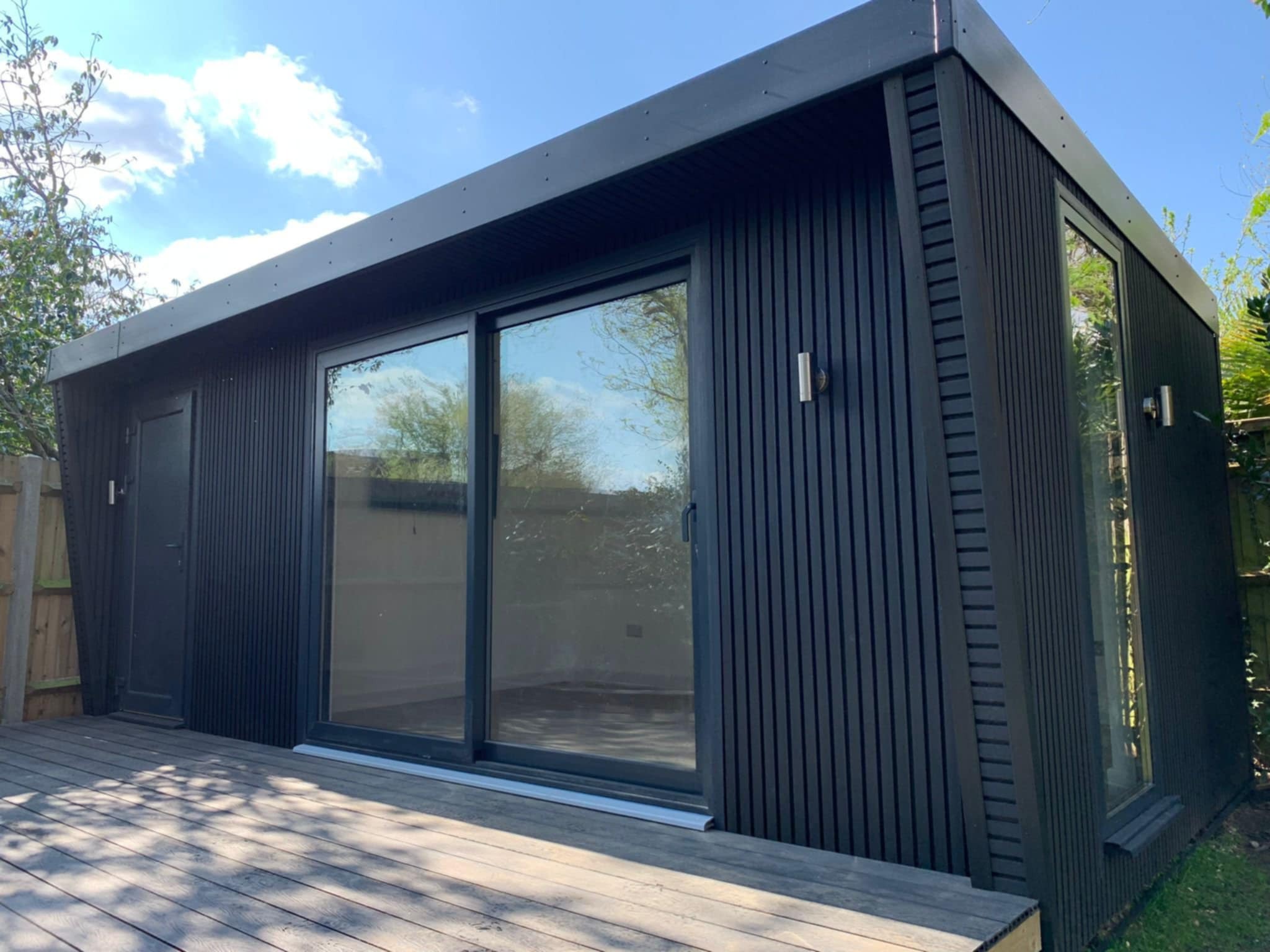 Nowoczesna, czarna altana z drewnianą elewacją i dużymi przesuwnymi drzwiami, osadzona na drewnianym tarasie w słoneczny dzień.