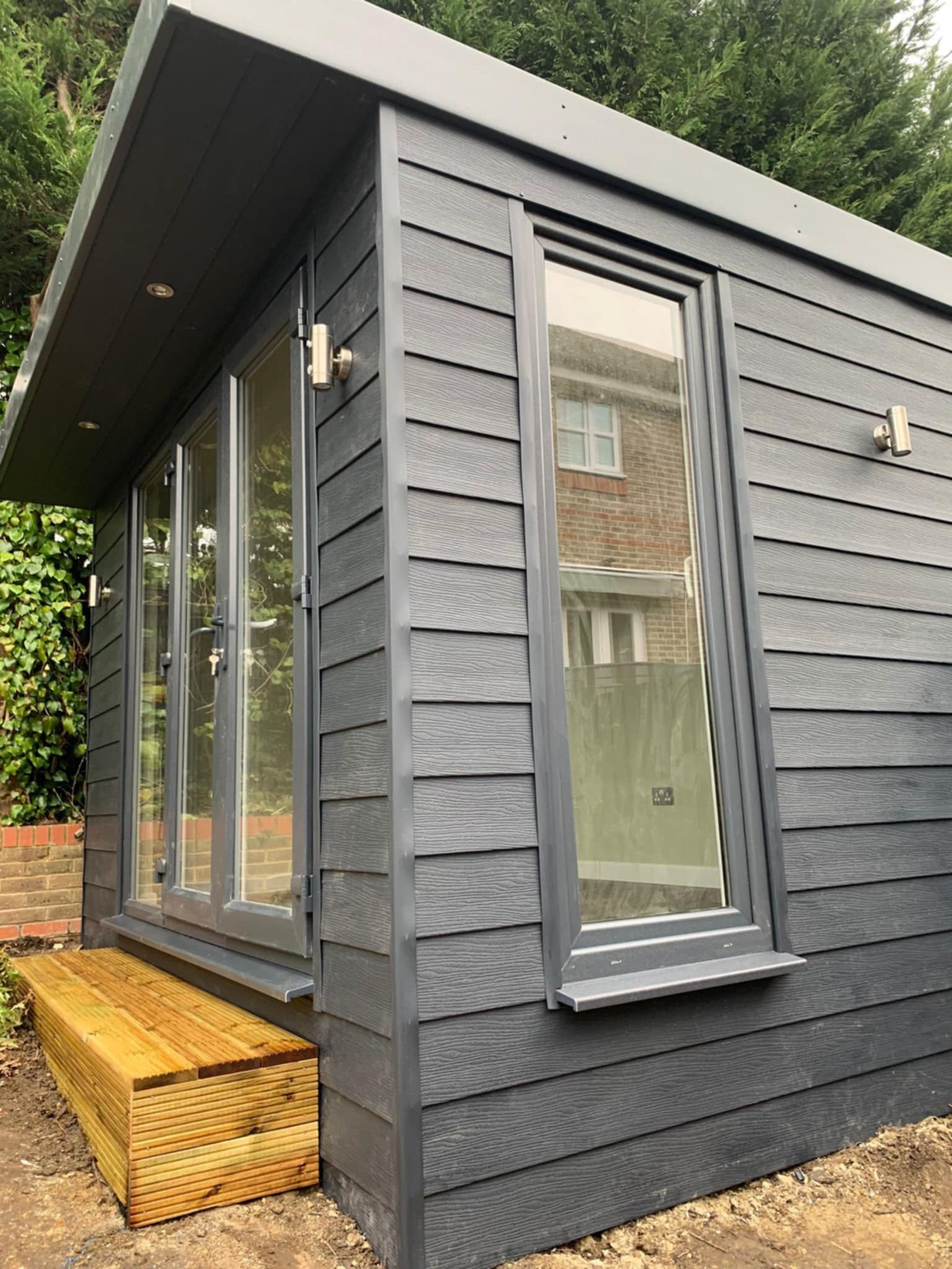 Nowoczesna, grafitowa altana z drewnianą podestem i dużymi oknami, otoczona zielenią. Widoczne oświetlenie zewnętrzne i struktura desek na elewacji.