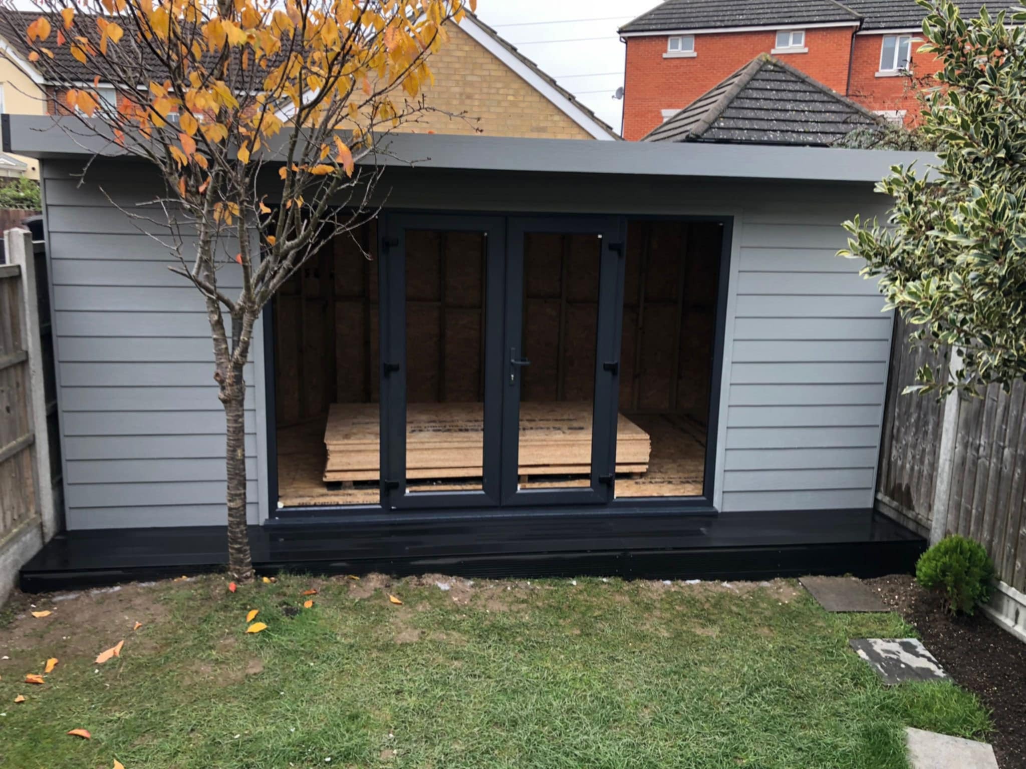 Nowoczesna, szara altana z dwuskrzydłowymi drzwiami, widok na wnętrze z ułożonymi płytami. Jesienne drzewo z żółtymi liśćmi dodaje uroku.