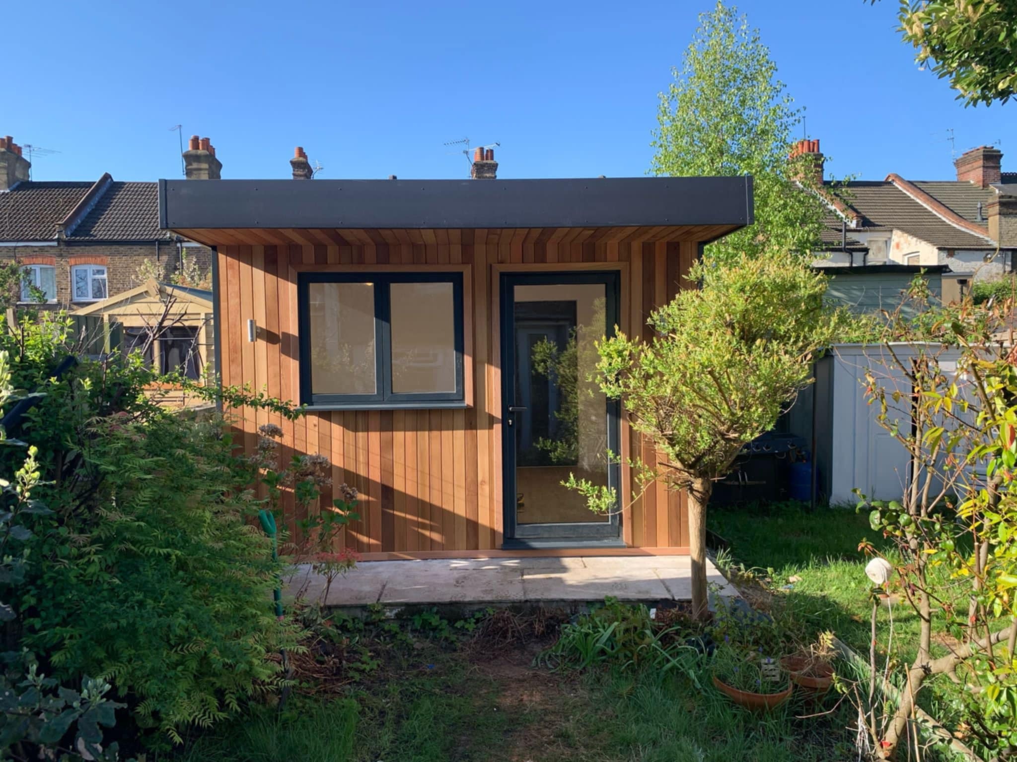 Nowoczesna altana z drewna w ogrodzie, z czarną ramą dachu i dużymi oknami, otoczona zielenią. Widoczne domy w tle.