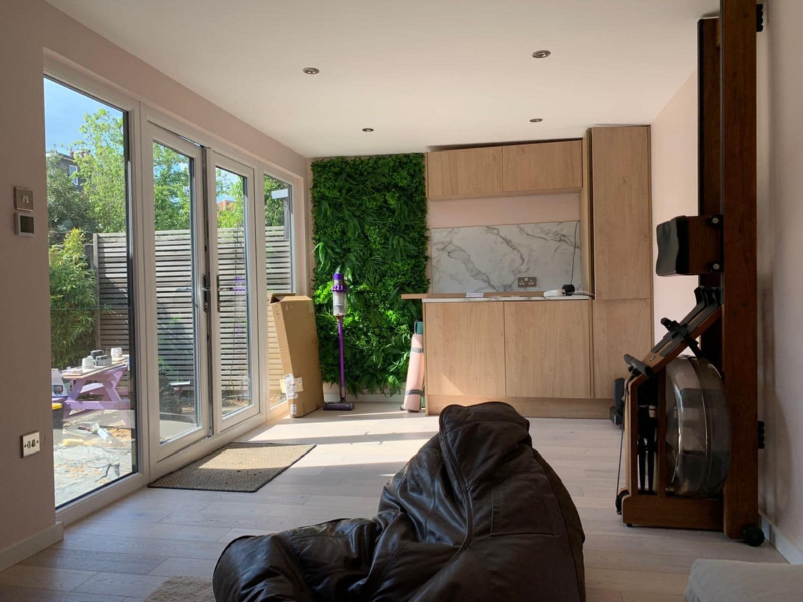 Jasne wnętrze z drewnianą zabudową kuchenną, ścianą z roślin, sprzętem do ćwiczeń i dużymi oknami balkonowymi. Widoczny fragment brązowego worka sako.