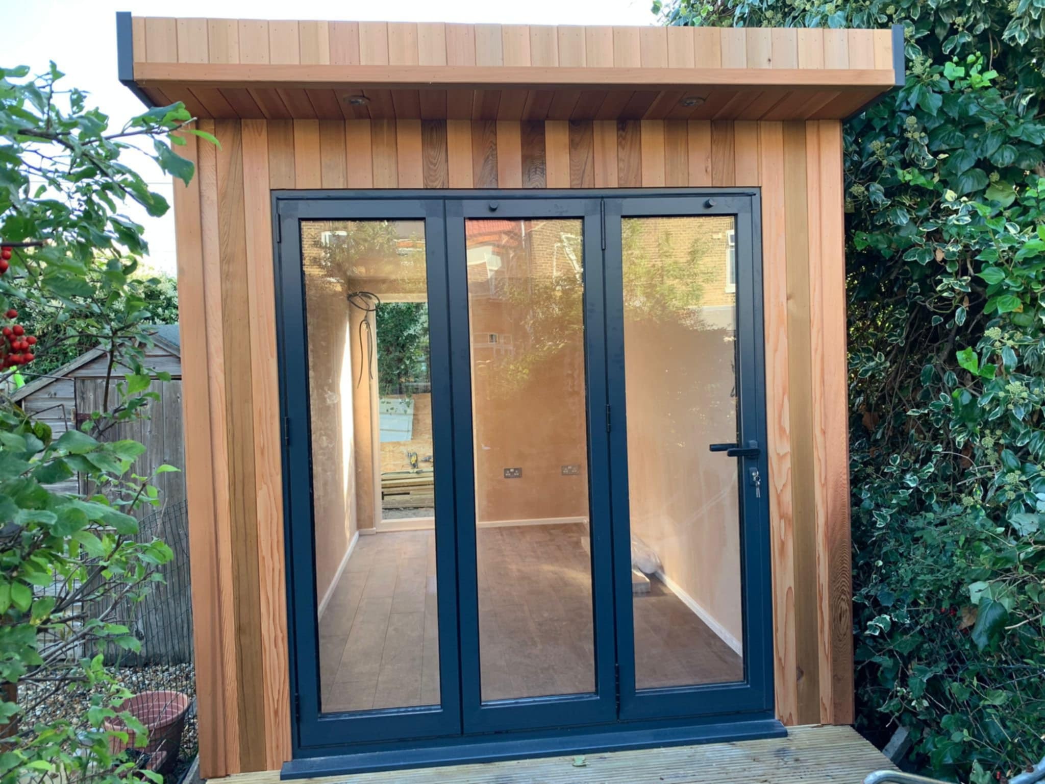 Nowoczesna, drewniana altana z trzyskrzydłowymi, przeszklonymi drzwiami w grafitowej ramie, z widocznym wnętrzem i elementami wyposażenia.