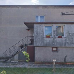 Pro-System Łukasz Kloskowski - Szary dom z balkonem i schodami, widoczne ślady wilgoci na elewacji, okna z kwiatami, fragment rusztowania po prawej stronie. Ujęcie z poziomu gruntu.