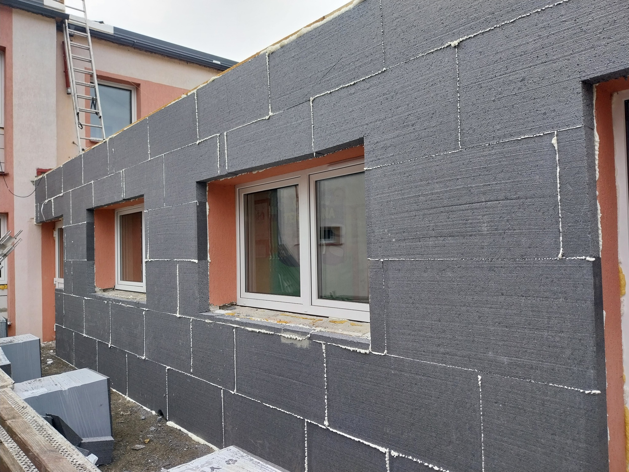 Ściana budynku w trakcie ocieplania szarym styropianem, widoczne okna i pianka uszczelniająca. Na drugim planie drabina oparta o budynek.