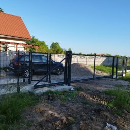 Dominik Domański - Nowoczesna, przesuwna brama panelowa w Żarowie, z czarną ramą i siatkowym wypełnieniem. W tle dom z czerwoną dachówką i zaparkowany samochód.