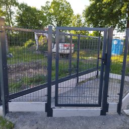 Dominik Domański - Nowoczesne, metalowe ogrodzenie panelowe z bramką, osadzone na betonowej podmurówce. W tle widoczny teren budowy z zaparkowaną koparką.