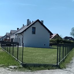 Dominik Domański - Nowoczesne, panelowe ogrodzenie w kolorze grafitowym okala zadbany trawnik przed nowo wybudowanym domem jednorodzinnym. Widok z boku, w tle kolejne domy.