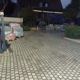Dominik Domański - Ułożona kostka granitowa na podjeździe. Widoczny fragment ogrodzenia, palety z materiałem budowlanym oraz dom w tle. Wykorzystano szarą kostkę o małym formacie.