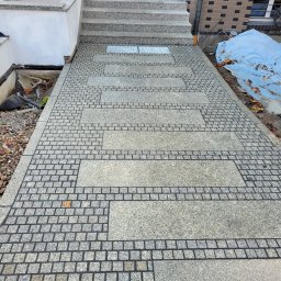 Dominik Domański - Granitowa ścieżka z dekoracyjnymi wzorami i schodami. Widoczny fragment budynku z oknem i rynną. Całość utrzymana w szarościach, podkreślona detalami.