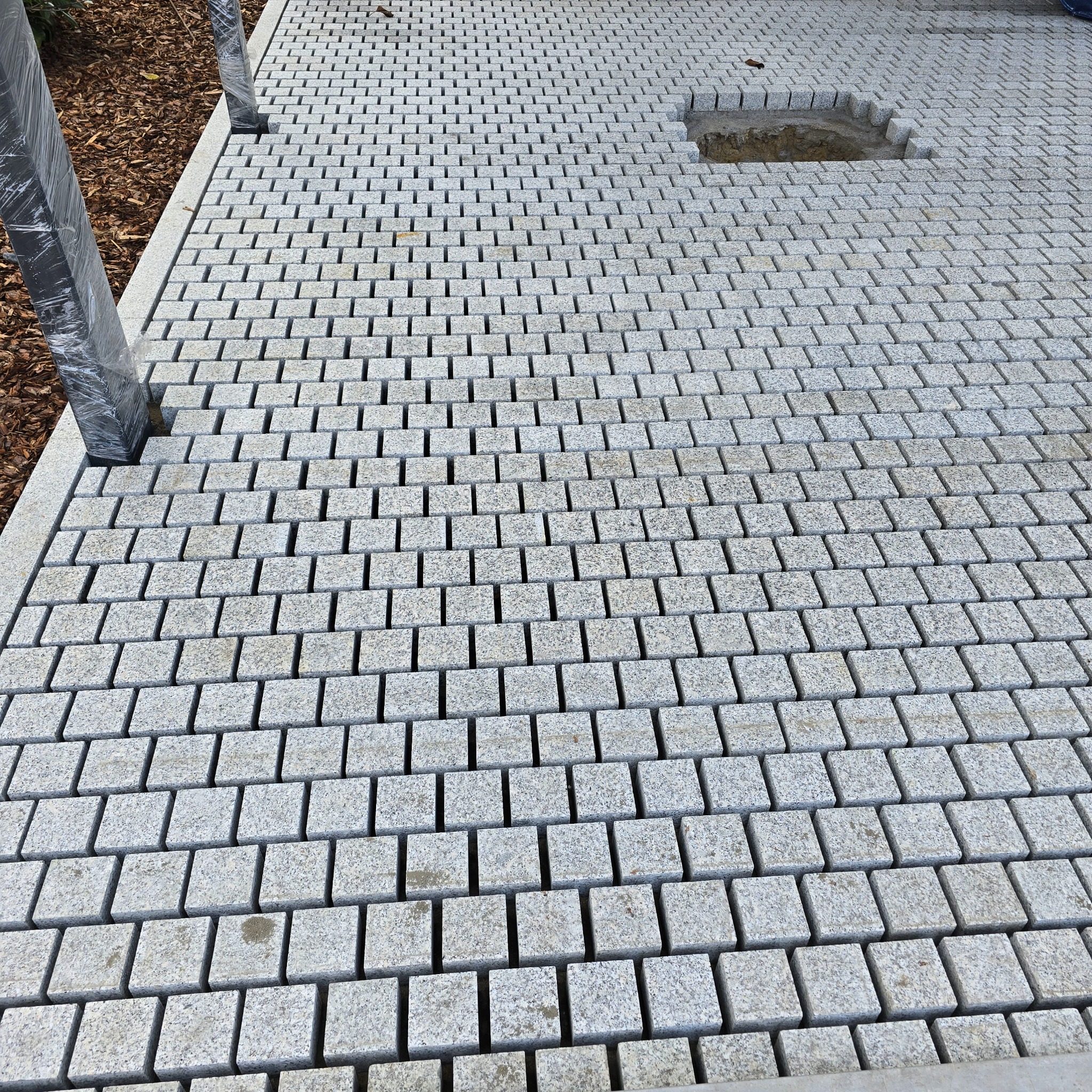 Ułożona szara kostka granitowa tworząca wzór. Fragment z wykopanym miejscem na roślinę. Elementy konstrukcyjne w folii ochronnej. Widok z góry.