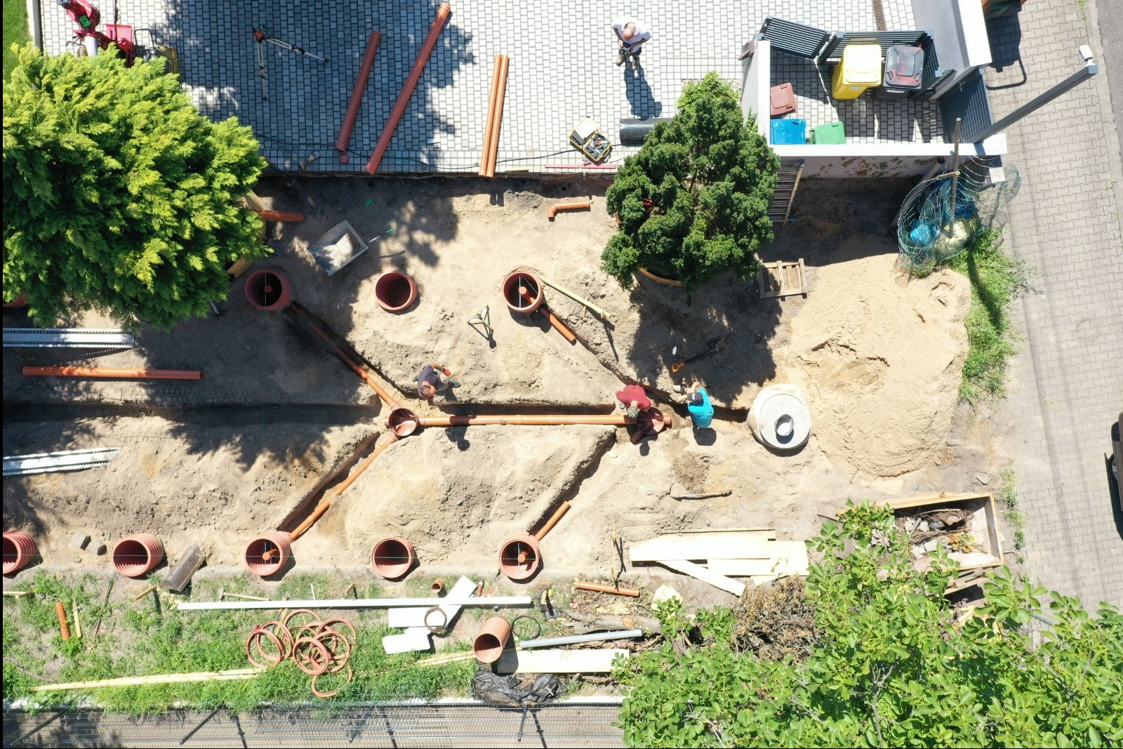 Z góry widok na montaż systemu drenażowego z rur PVC w wykopie. Trzech pracowników łączy rury, widoczne studzienki rewizyjne, narzędzia i częściowo ułożona kostka.