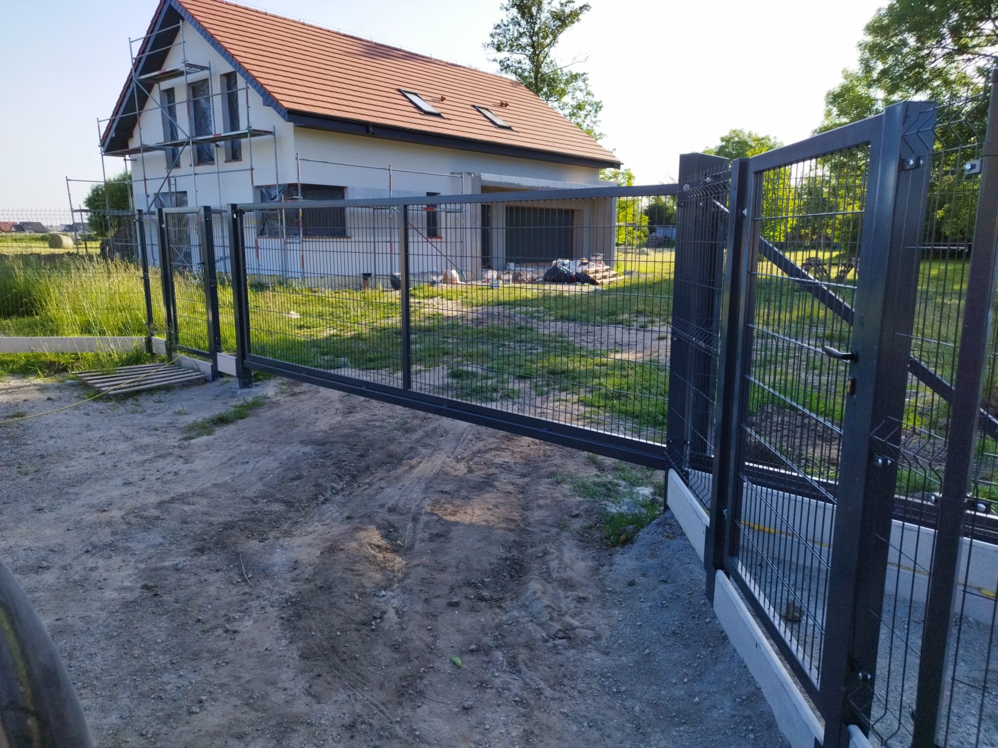 Nowoczesne ogrodzenie panelowe z metalową bramą przesuwną na tle domu w budowie. Widoczne betonowe podmurówki i trawnik. Ujęcie z bliska.
