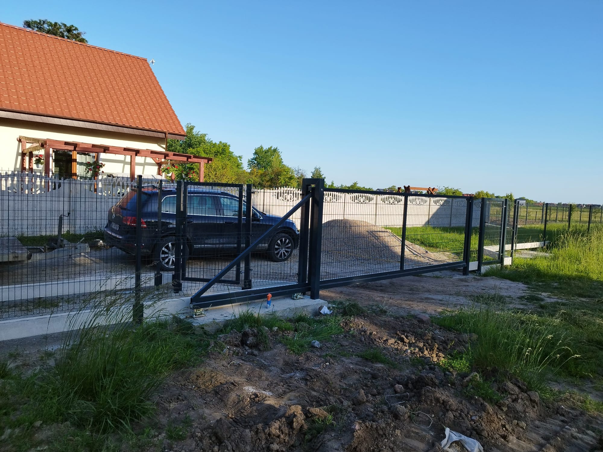 Nowoczesna, przesuwna brama panelowa w Żarowie, z czarną ramą i siatkowym wypełnieniem. W tle dom z czerwoną dachówką i zaparkowany samochód.