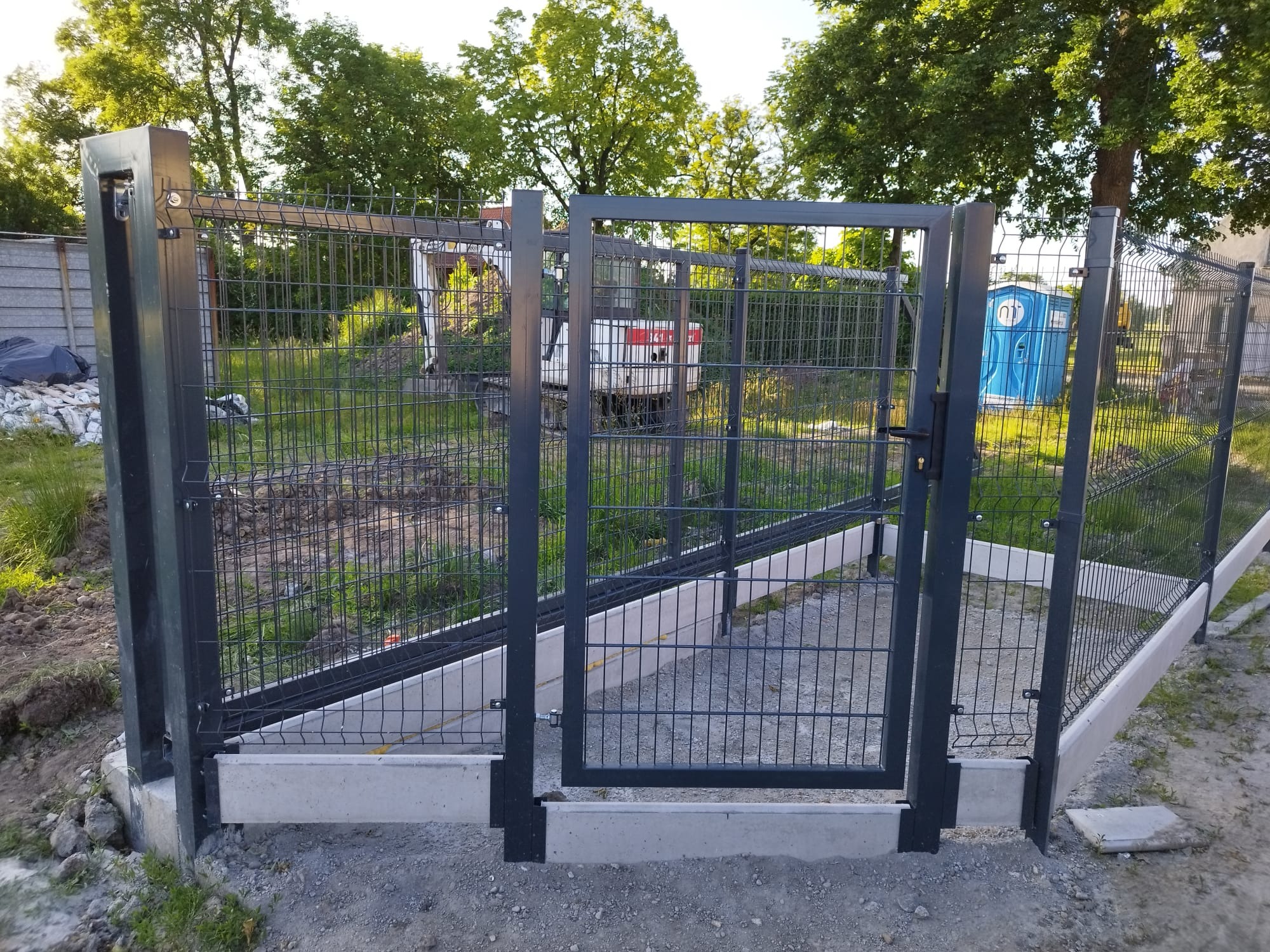 Nowoczesne, metalowe ogrodzenie panelowe z bramką, osadzone na betonowej podmurówce. W tle widoczny teren budowy z zaparkowaną koparką.