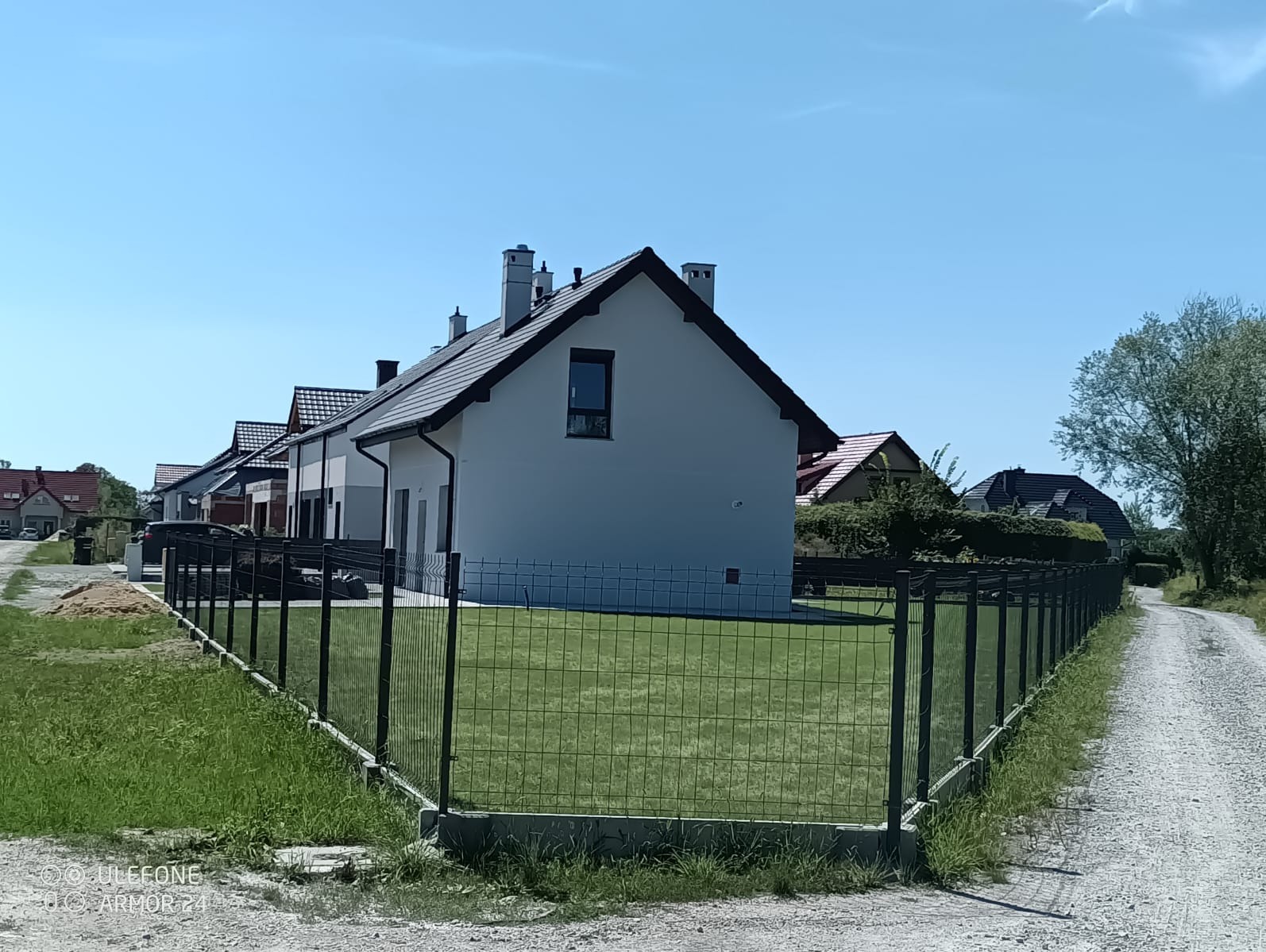 Nowoczesne, panelowe ogrodzenie w kolorze grafitowym okala zadbany trawnik przed nowo wybudowanym domem jednorodzinnym. Widok z boku, w tle kolejne domy.