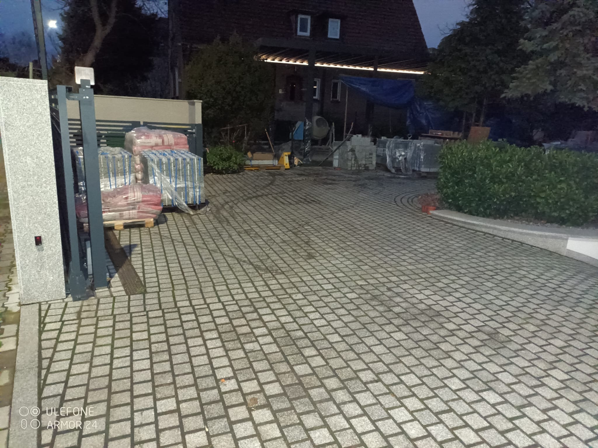 Ułożona kostka granitowa na podjeździe. Widoczny fragment ogrodzenia, palety z materiałem budowlanym oraz dom w tle. Wykorzystano szarą kostkę o małym formacie.