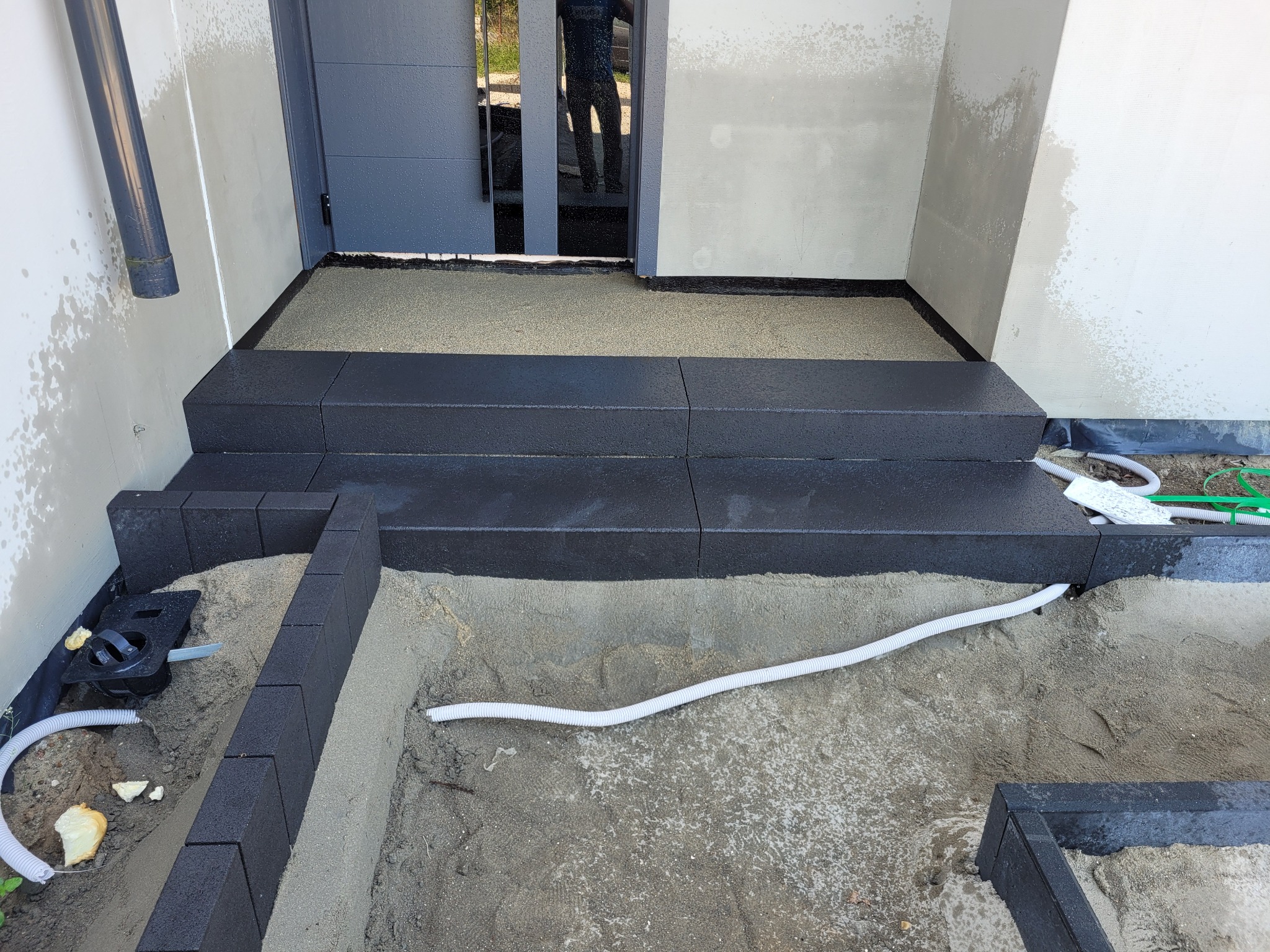 Ciemne, prostokątne stopnie wejściowe z kostki, częściowo ułożone, z widocznymi rurami drenażowymi i piaskiem. Wejście do domu w nowoczesnym stylu.