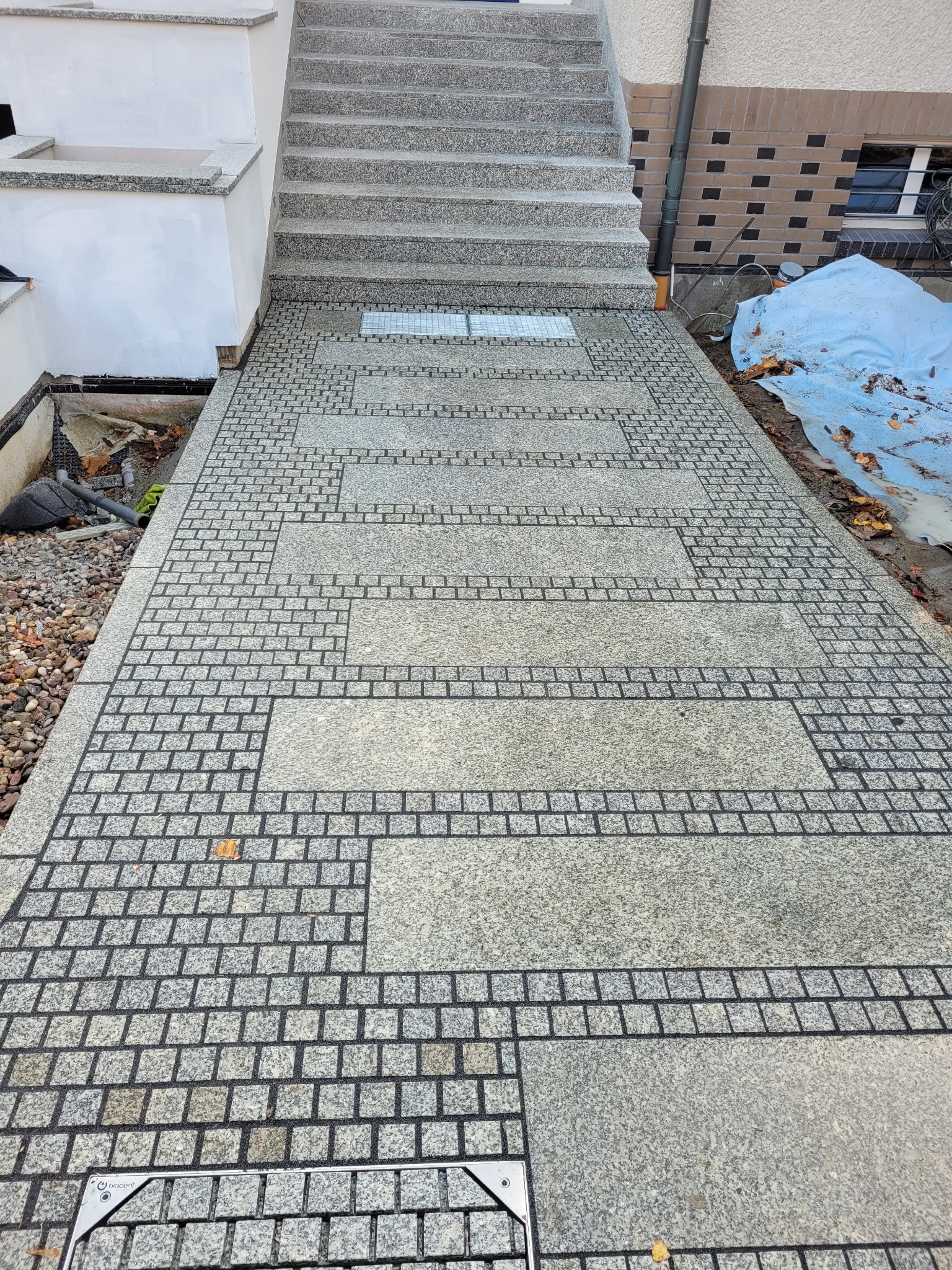 Granitowa ścieżka z dekoracyjnymi wzorami i schodami. Widoczny fragment budynku z oknem i rynną. Całość utrzymana w szarościach, podkreślona detalami.