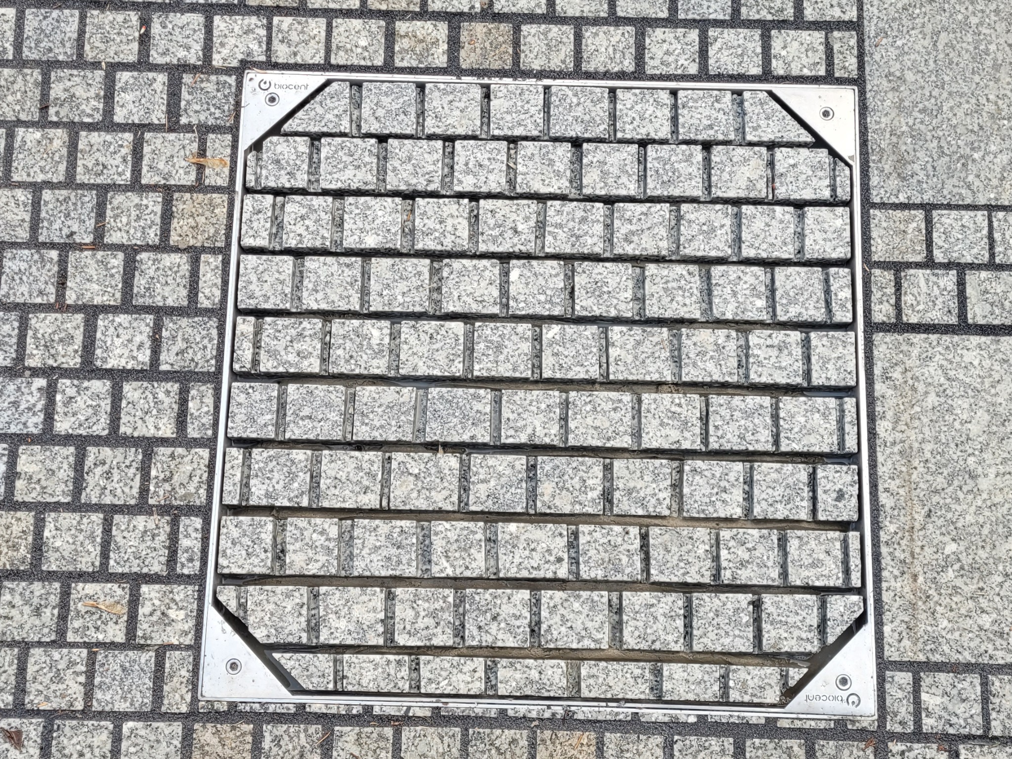 Szara kostka granitowa ułożona wokół metalowej kratki ściekowej z logo, tworząca równą, estetyczną powierzchnię. Widok z góry.