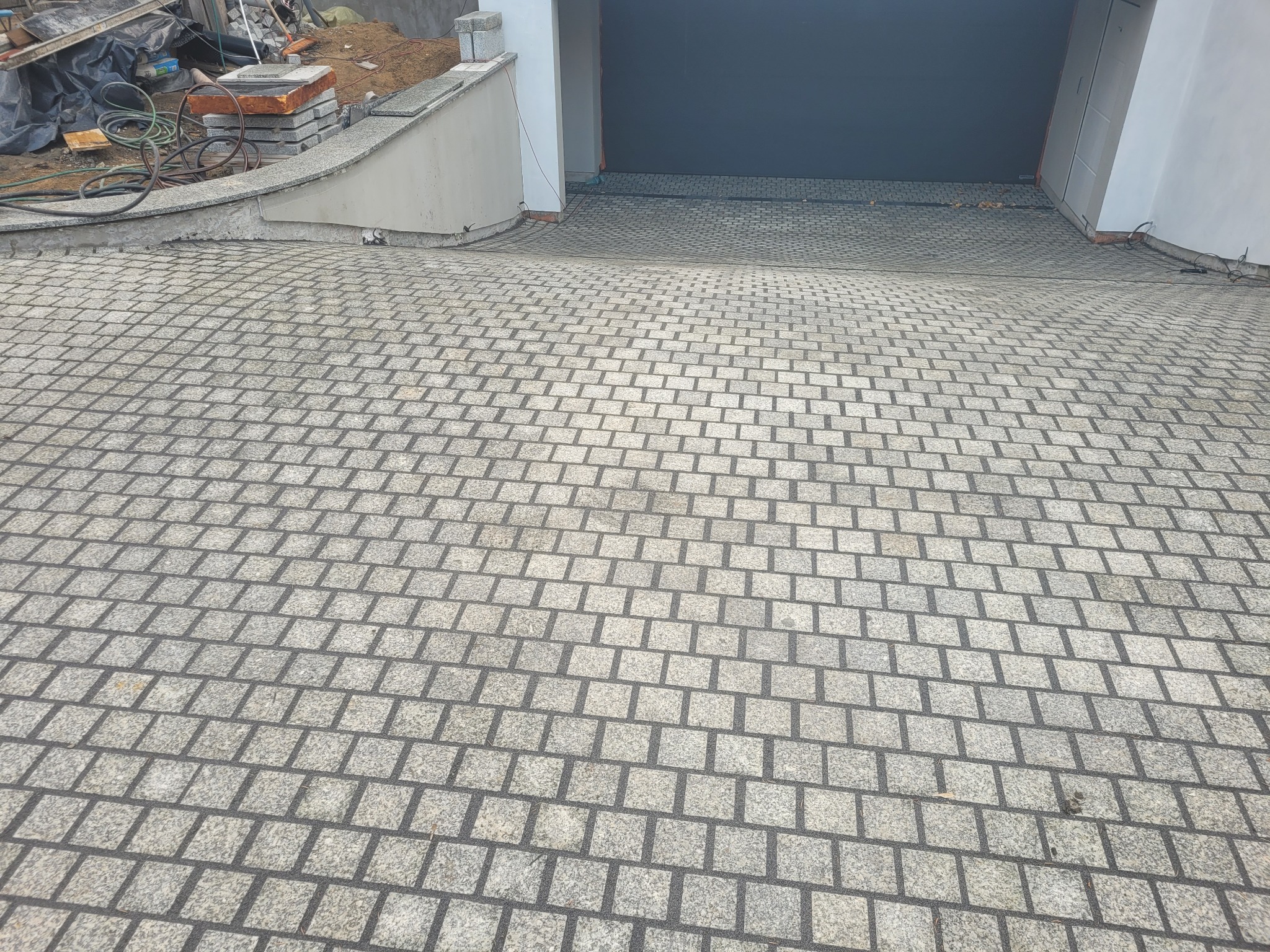 Szara kostka granitowa ułożona na podjeździe do garażu, widok z góry. Delikatny spadek terenu. Fragment murku i brama garażowa w tle.