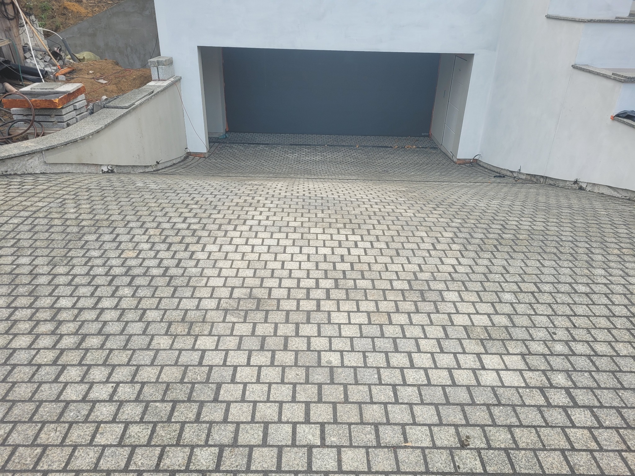 Ułożona kostka granitowa w szarym odcieniu tworzy podjazd do garażu, widoczny fragment posesji w tle. Regularny wzór kostki.