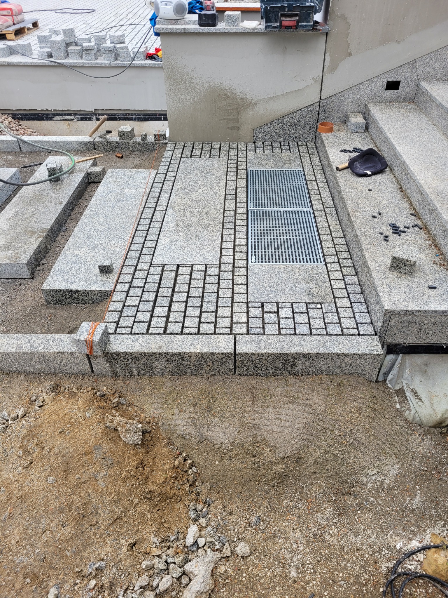 Szara kostka granitowa ułożona w regularny wzór z kratką drenażową. Widoczne niedokończone elementy i narzędzia, co sugeruje etap pracy.
