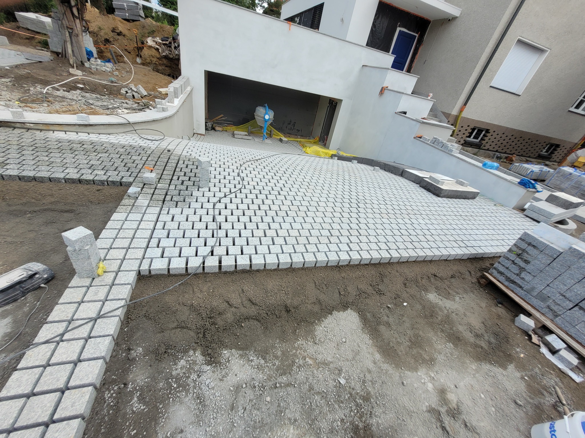 Układanie kostki granitowej na podjeździe do garażu. Częściowo ułożona kostka w regularny wzór, widoczne zapasy kostki i przygotowane podłoże. Prace w toku.