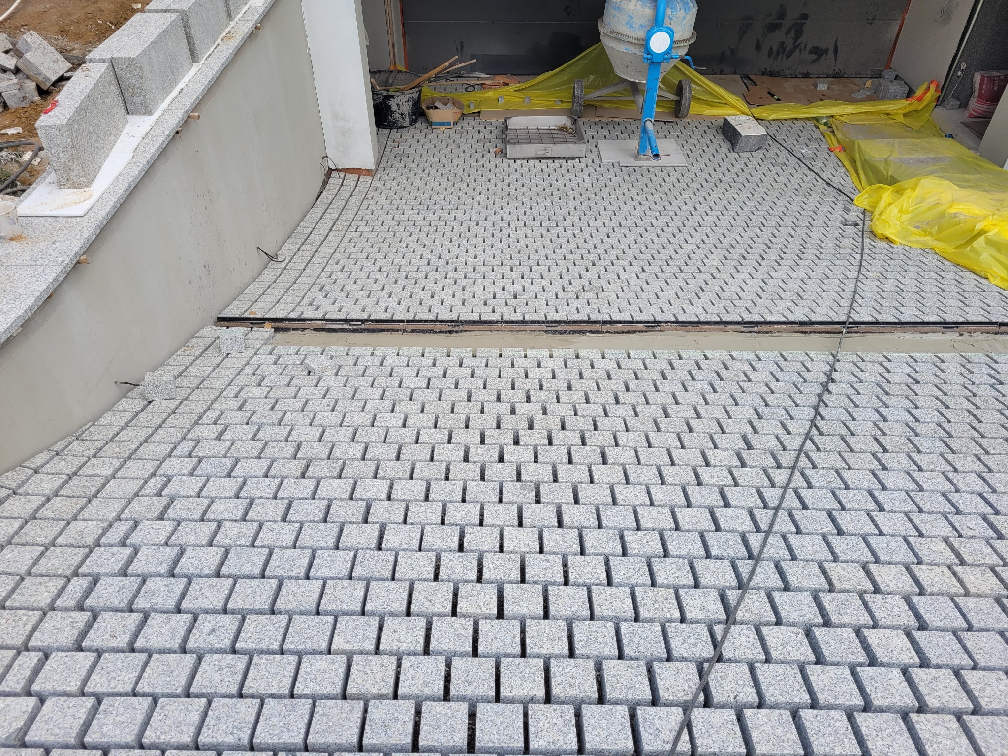 Jasnoszara kostka granitowa ułożona w regularny wzór, widoczny fragment betoniarki, materiały budowlane w tle. Krawężnik z kostki wzdłuż ściany.