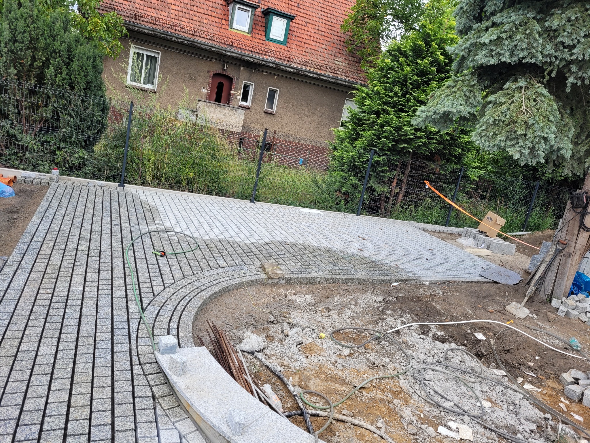 Fragment układania szarej kostki granitowej na podjeździe z zaokrąglonym brzegiem, widoczny teren budowy i dom w tle.