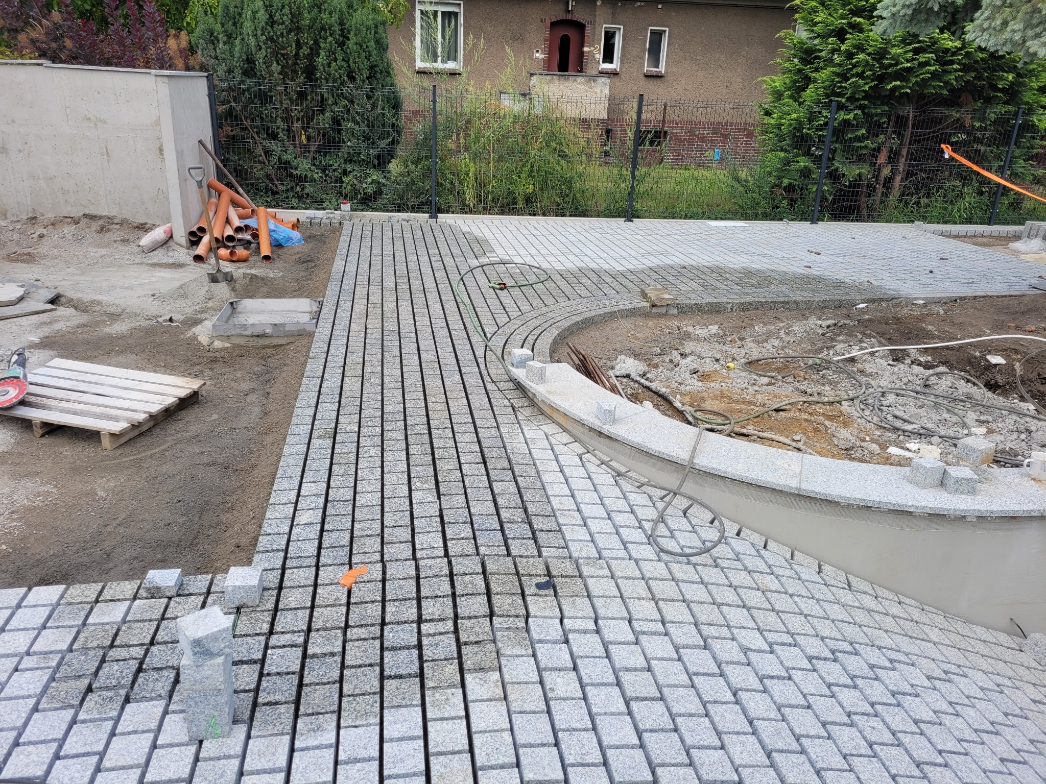 Fragment układania szarej kostki granitowej w regularny wzór. Częściowo ułożony chodnik z obrzeżem. Na drugim planie rury drenażowe i budynek z cegły.