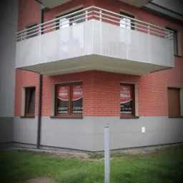 Czerwony budynek z balkonem i oknami z naklejkami. Widok z dołu, trawnik przed budynkiem. Firma 'Koala' na naklejkach w oknach.