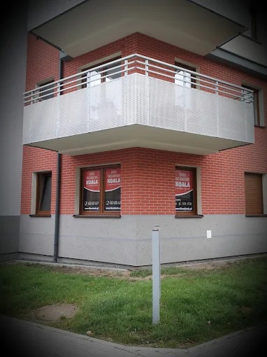 Czerwony budynek z balkonem i oknami z naklejkami. Widok z dołu, trawnik przed budynkiem. Firma 'Koala' na naklejkach w oknach.