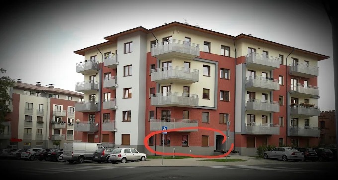 Budynek mieszkalny z biurem na parterze, widoczne zaparkowane samochody i chodnik. Lokalizacja w mieście.