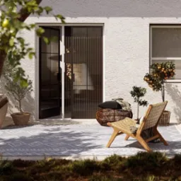 Balkon z roletą plisowaną w czarnej ramie, otwartą na taras z krzesłem i roślinami. Elegancki design z widoczną fakturą ściany i wzorem dywanu.